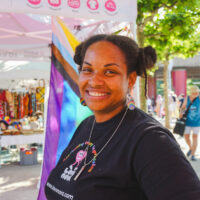 Smiling person with braided hair buns and colorful earrings in a black T-shirt at an outdoor Pride market